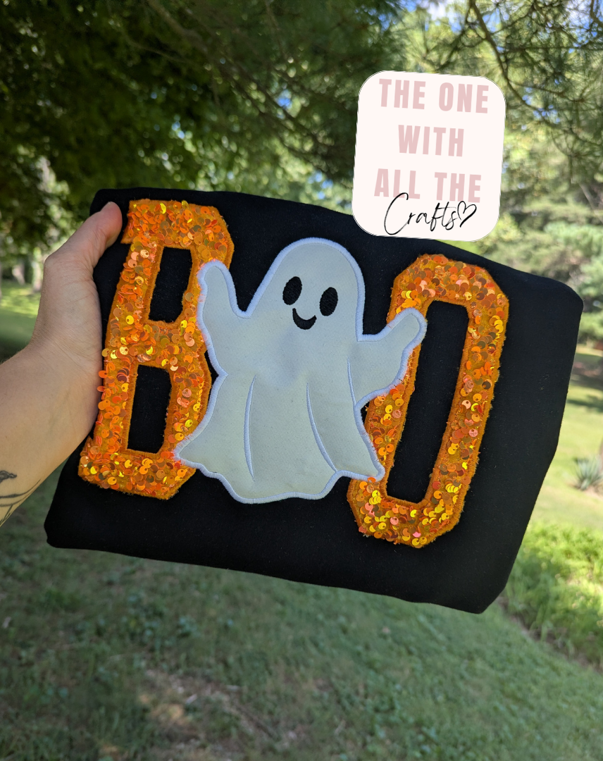A hand holds a black fabric sign spelling BOO with orange sequined letters and a white ghost in the center, photographed outdoors with trees and grass in the background. A pink sticker reading 'THE ONE WITH ALL THE Crafts' is attached near the top.