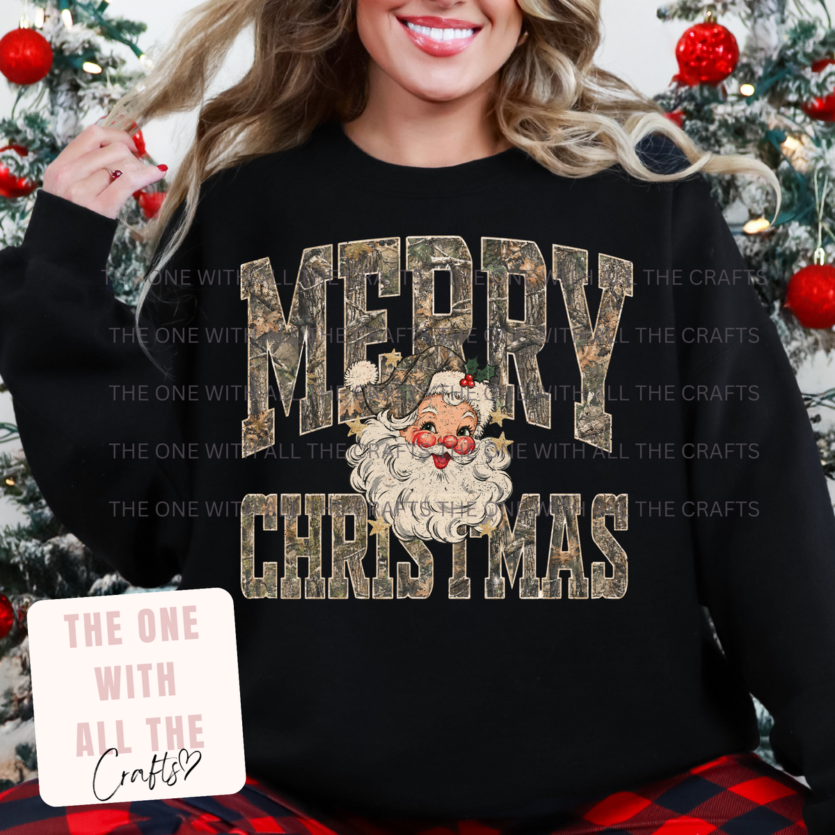 A woman wearing a black crewneck sweatshirt with a 'MERRY CHRISTMAS' graphic featuring a camouflage-textured font and a Santa Claus face in the center. The background shows a decorated Christmas tree.