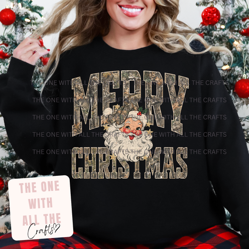 A woman wearing a black crewneck sweatshirt with a 'MERRY CHRISTMAS' graphic featuring a camouflage-textured font and a Santa Claus face in the center. The background shows a decorated Christmas tree.