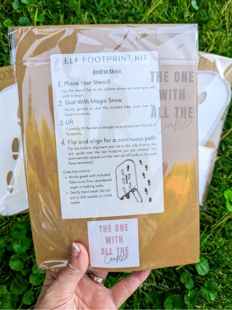 A hand holds a clear plastic bag containing an Elf Footprint Kit outdoors on green grass. The package includes printed instructions on a white label and a brown paper insert.