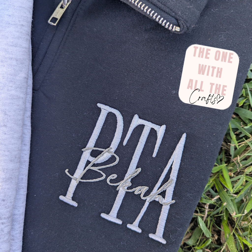 Close-up of a dark navy sweatshirt or hoodie showing embroidered text 'PTA Bekah' in light gray and white, a zipper, and a pink-and-white sticker that reads 'THE ONE WITH ALL THE Crafts♡'. Grass and part of a light gray garment are visible around it.