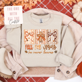 A beige crewneck sweatshirt laid flat with a folded light pink cardigan, autumn leaves, pumpkins, and a red plaid blanket in the background. The sweatshirt has a graphic of three striped gift boxes with bows and the text 'FALL FOR JESUS he never leaves'.