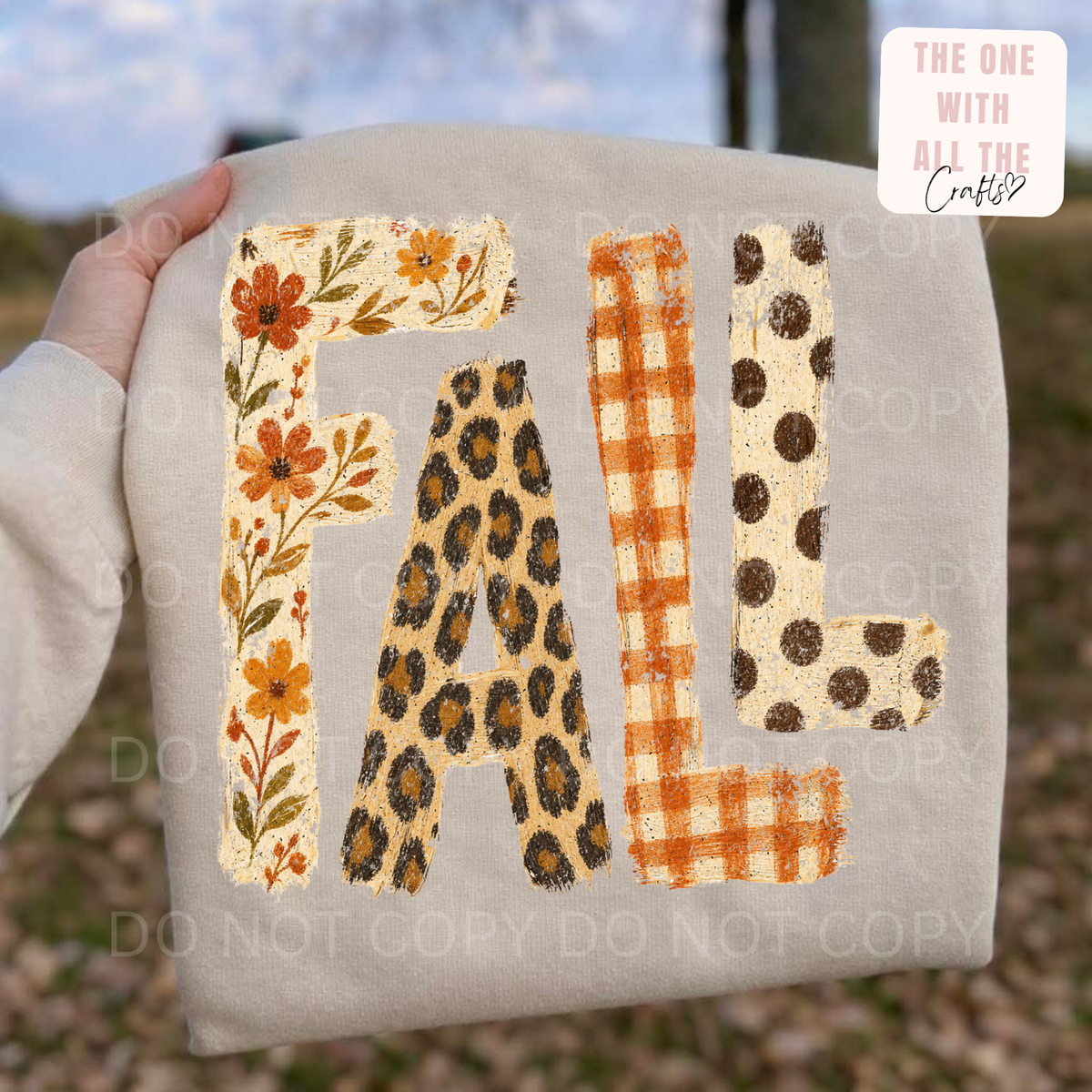 A hand holds a beige fabric pillow or cushion outdoors, featuring the word "FALL" in decorative patchwork-style letters with floral, leopard print, gingham, and polka-dot patterns. A small logo in the top right reads "THE ONE WITH ALL THE Crafts," and faint watermark text appears across the image.