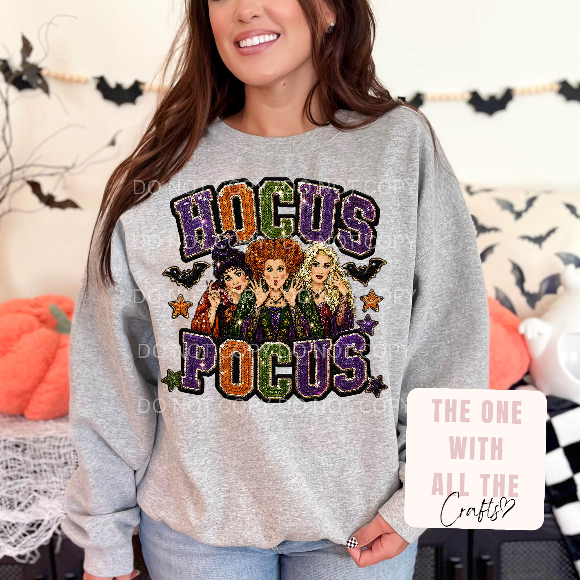 A woman wearing a light gray heather crewneck sweatshirt with a colorful 'Hocus Pocus' graphic featuring the three Sanderson sisters and stars. The background shows a cozy, decorated room with Halloween elements like pumpkins and black bat cutouts.