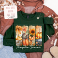A dark green long-sleeve t-shirt featuring a vertical autumn-themed graphic print with pumpkins, sunflowers, and fall foliage, styled in a flat-lay arrangement with a brown hoodie, jeans, and a decorative card.