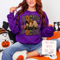 A woman wearing a purple crewneck sweatshirt with a 'Hocus Pocus' graphic featuring Sanderson, Wicked, and Mary Sue, sitting on an orange and white cushion with a mug.
