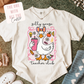 A cream-colored graphic t-shirt laid flat on a textured green carpet, featuring a cute illustration of a goose in a pink bow holding a pink drink, with the words 'silly goose teacher club'. A white sticker overlay in the top left reads 'THE ONE WITH ALL THE Crafty'.