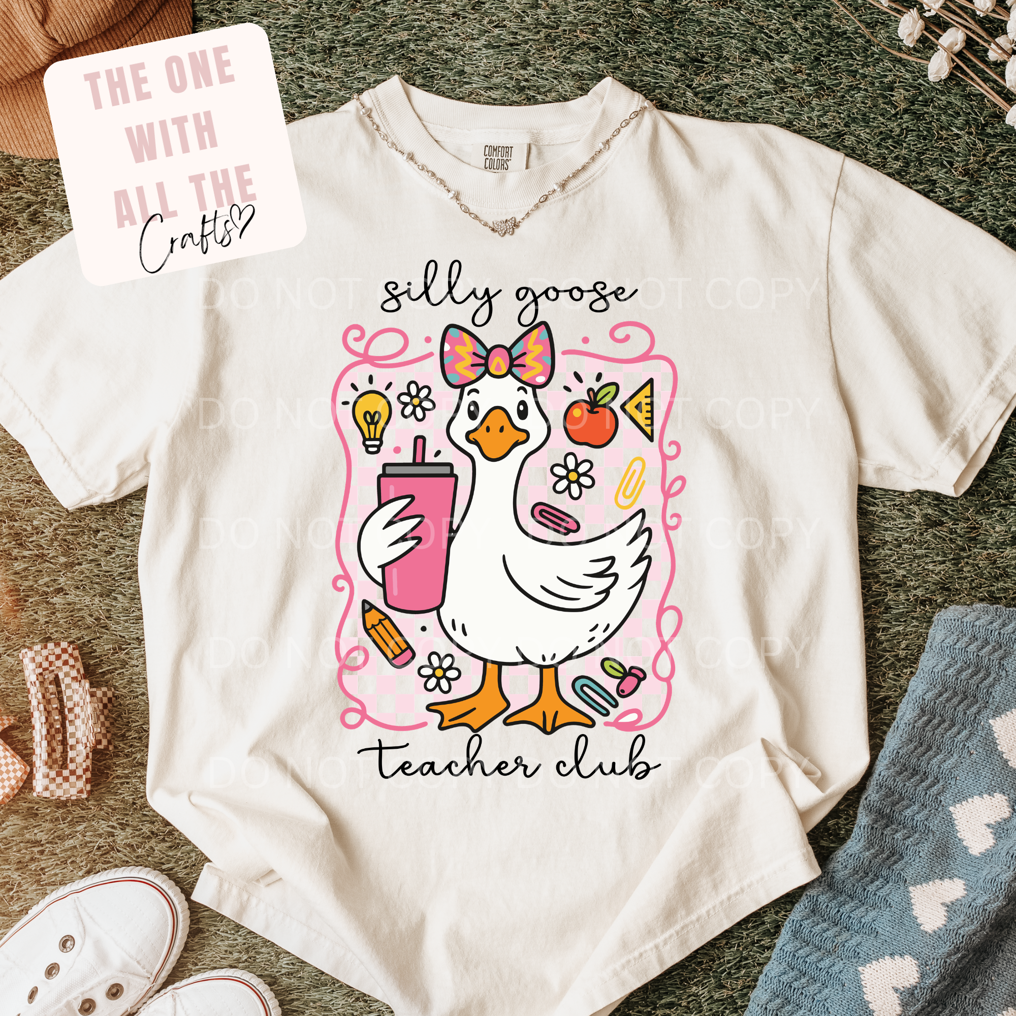 A cream-colored graphic t-shirt laid flat on a textured green carpet, featuring a cute illustration of a goose in a pink bow holding a pink drink, with the words 'silly goose teacher club'. A white sticker overlay in the top left reads 'THE ONE WITH ALL THE Crafty'.