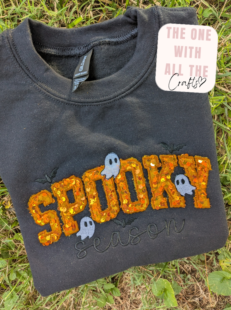 A folded dark charcoal gray sweatshirt laid on grass, featuring a glittery orange 'SPOOKY' graphic with small ghost details and the words 'season' underneath. A white sticker tag reading 'THE ONE WITH ALL THE Crafts' is attached near the collar.