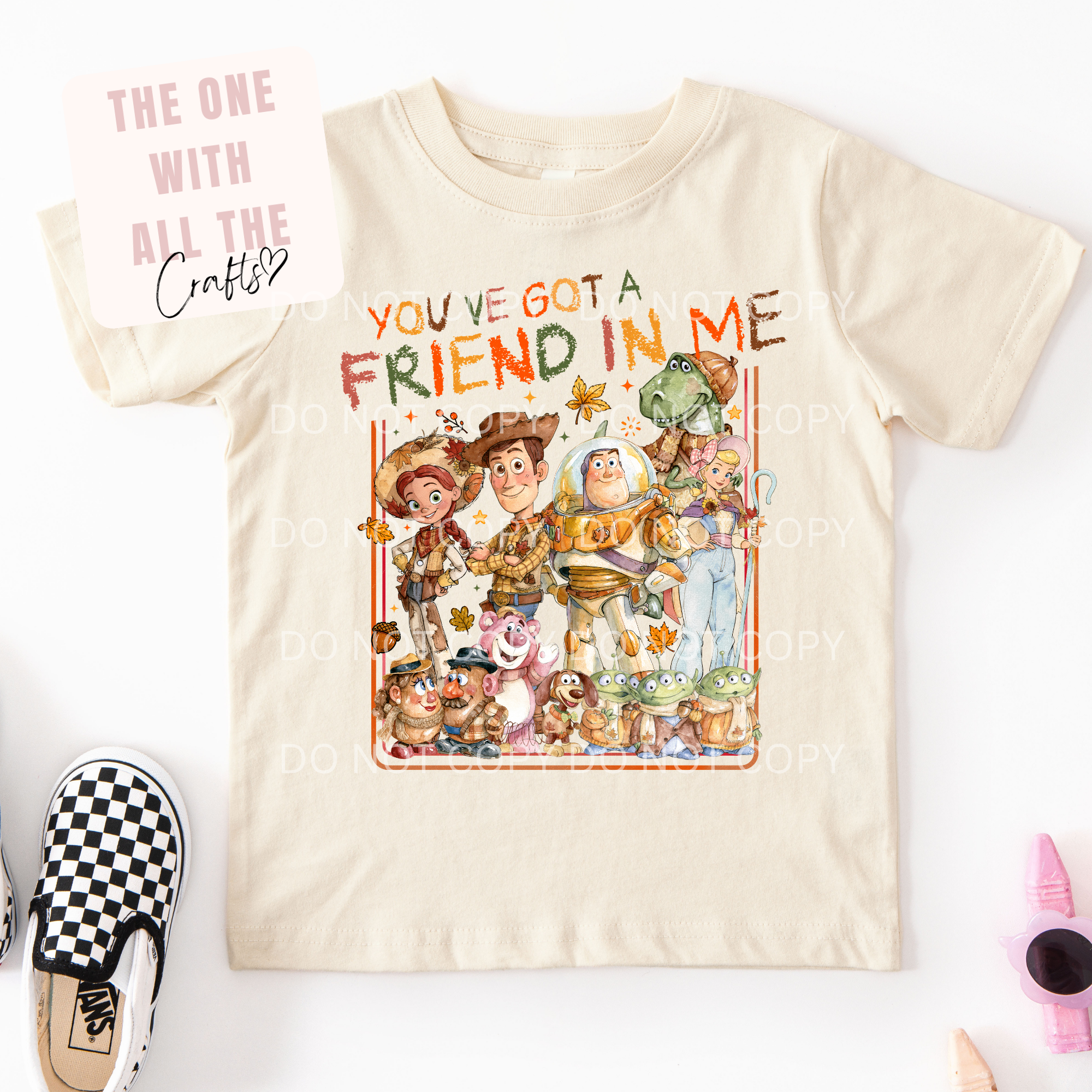A cream-colored kids' t-shirt featuring a colorful graphic of Toy Story characters with the text 'YOU'VE GOT A FRIEND IN ME'. The shirt is displayed flat on a white background with a shoe and small accessories as props.